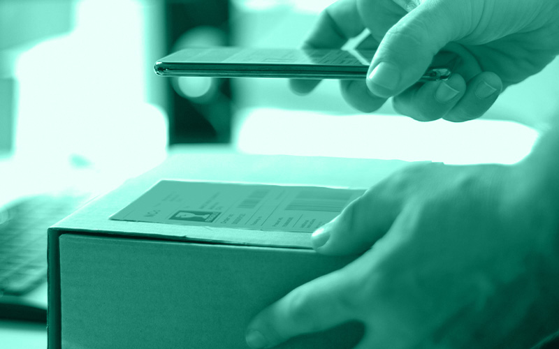 auf einem Karton bzw. einer Verpackung prüft ein Mensch mit seinem Smartphone Codes und Klarschrift auf einem Etikett