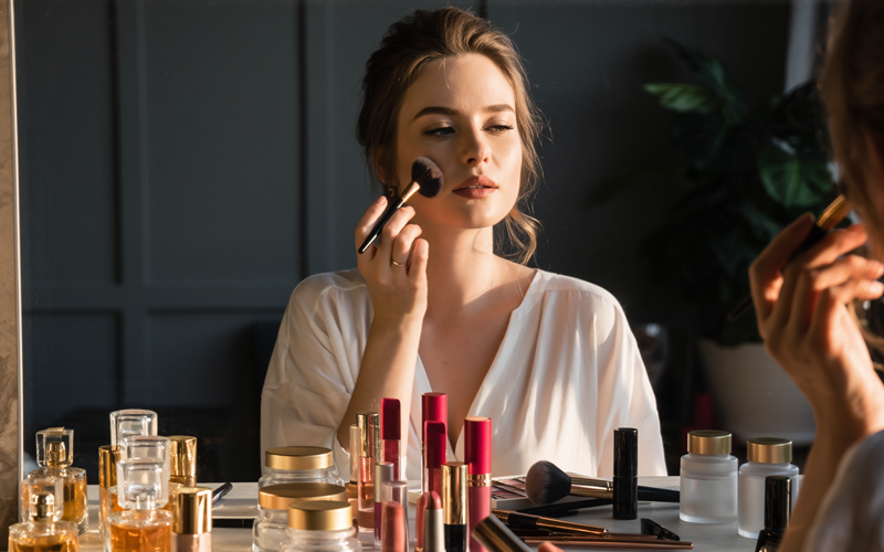 A woman sits in front of a mirror and applies makeup.